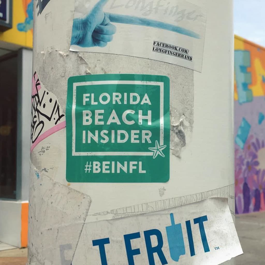 A photo of a streetlight pole with a teal Florida Beach Insider sticker stuck to it. Behind the sticker, there are all kinds of other stickers.